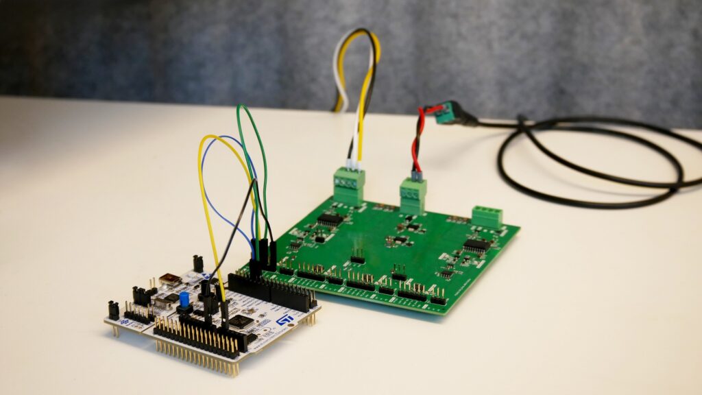 Gezeigt wird eine grüne Leiterplatte (PCB) mit verschiedenen Bauteilen, verbunden mit einem Entwicklungsboard. Farbige Kabel (gelb, blau, schwarz und grün) und Steckverbindungen verdeutlichen die Hardware-Interaktion. Im Hintergrund ist ein USB-Kabel sichtbar, das die Verbindung zur Stromversorgung oder Datenübertragung andeutet. Die Anordnung symbolisiert praktische Entwicklungs- und Testprozesse. Es steht für die Elektronikentwicklung.