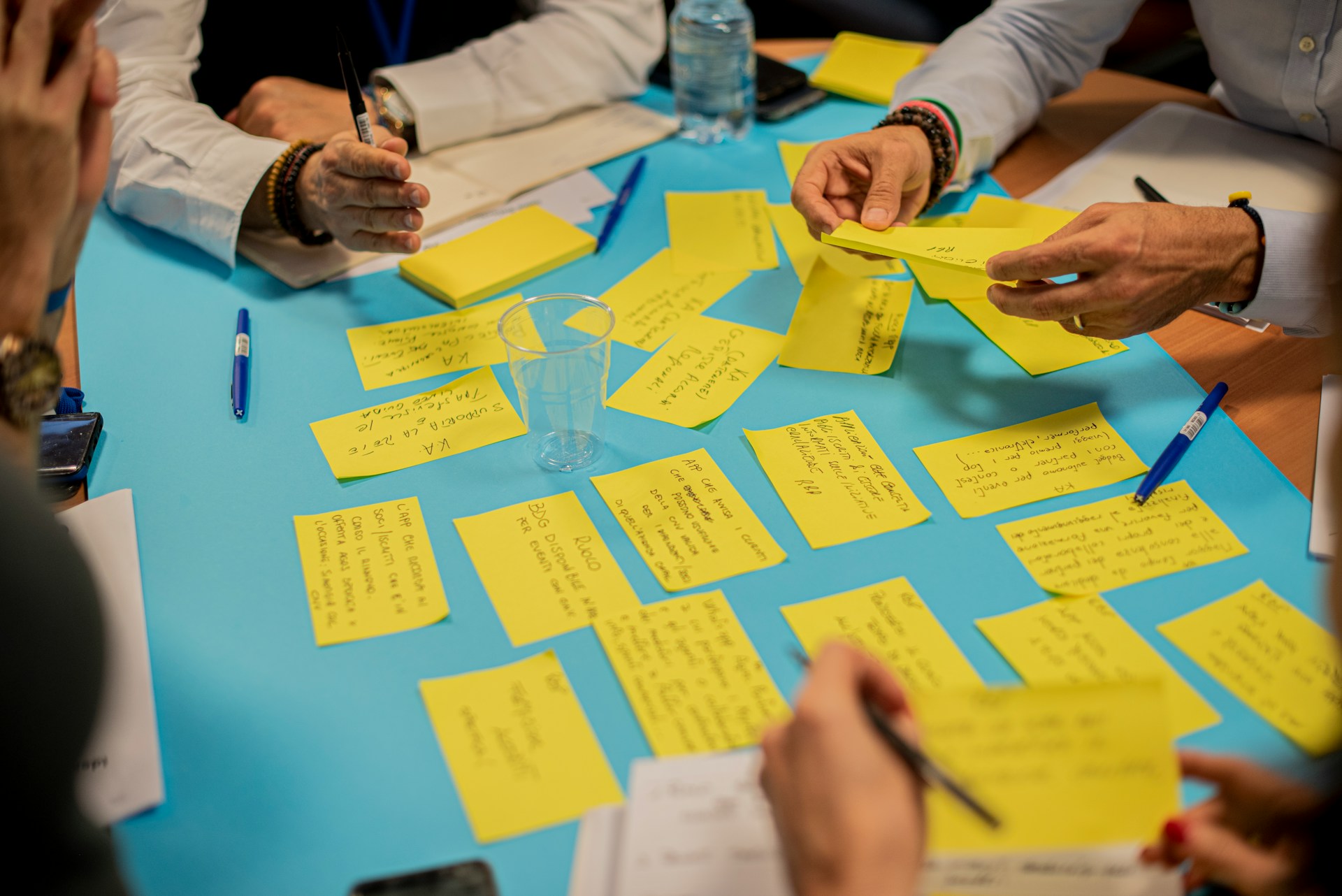 Personen arbeiten gemeinsam an einem Anforderungsworkshop, indem sie Ideen auf gelben Haftnotizen sammeln und sortieren. Ein Tisch mit blauer Unterlage ist mit zahlreichen Notizzetteln bedeckt, während Teilnehmer mit Stiften und Notizbüchern arbeiten.